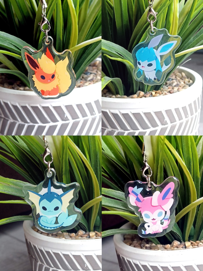 May include: Four clear acrylic charms featuring different Pokemon characters. The charms are attached to silver chains. The charms are displayed against a background of green plants in white pots.