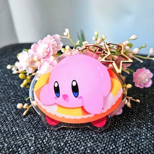 May include: Pink and yellow character keychain with a star charm. The character is a pink creature with big eyes and a small mouth. The keychain is made of clear acrylic.
