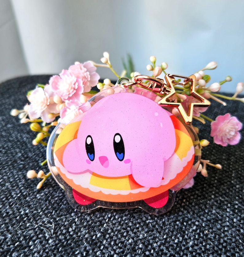 May include: Pink and yellow character keychain with a star charm. The character is a pink creature with big eyes and a small mouth. The keychain is made of clear acrylic.