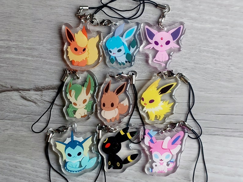 May include: A set of ten clear acrylic charms featuring different Pokemon characters. The charms are attached to black string lanyards. The characters include Eevee, Flareon, Jolteon, Vaporeon, Espeon, Umbreon, Leafeon, Glaceon, Sylveon, and a Pikachu.