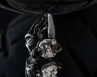 May include: A silver and black steampunk-style pendant with intricate gears and a detailed design. The pendant is hanging from a black chain.