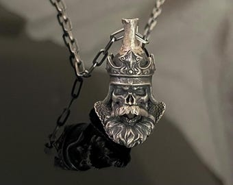 May include: Silver pendant necklace featuring a detailed skull with a crown and beard. The pendant is attached to a silver chain.