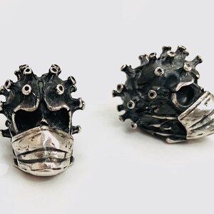 May include: A pair of silver skull earrings with a textured surface and a face mask. The skulls have a spiky, corona-like design.