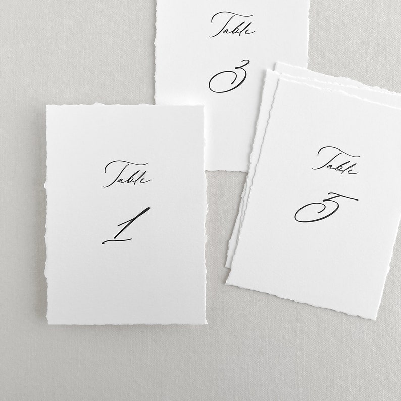 Pu&ograve; includere: Tre carte di numero di tavolo bianche con calligrafia nera. Le carte sono numerate 1, 2 e 3. Le carte hanno un bordo ruvido e testurizzato.