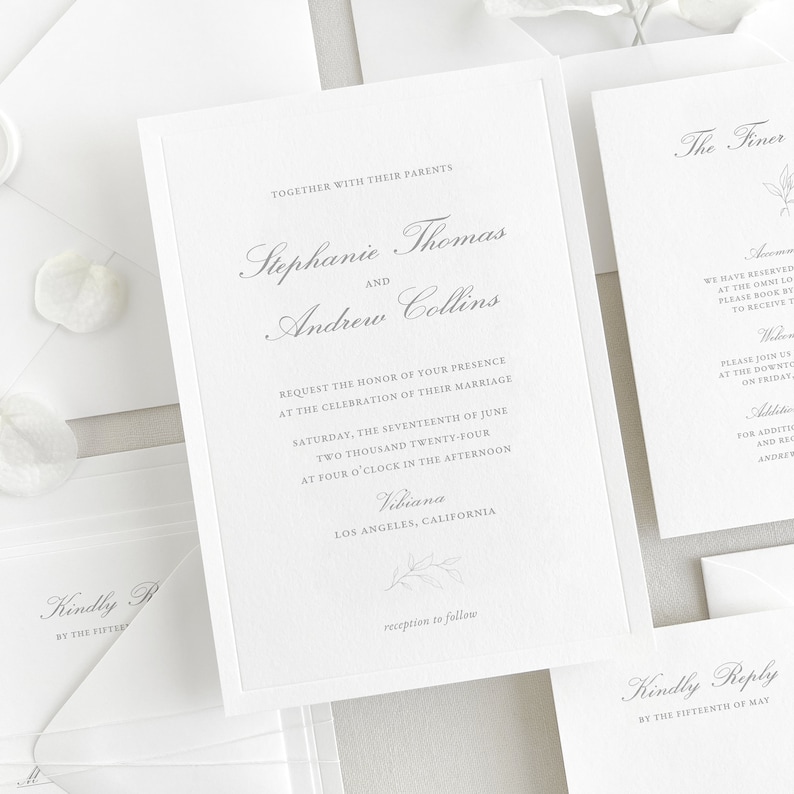 Invitation card features a thin de-bossed border, a classic script font for the names/venue, and a classic block font for the main text. A hand-drawn floral sprig is placed at the bottom of the card. Shown with matching white envelopes.