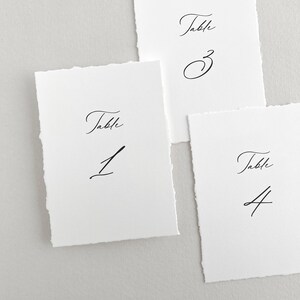 Pu&ograve; includere: Tre carte di numero di tavolo bianche con calligrafia nera. Le carte sono numerate 1, 3 e 4. Ogni carta ha la parola "Tavolo" scritta sopra il numero.