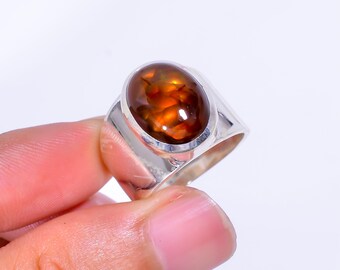 May include: A silver ring featuring a large, oval-shaped cabochon gemstone with a warm, amber-brown color. The gemstone has a polished, glossy surface. The ring's band is a simple, modern design.