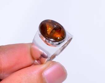 Op de afbeelding: Zilveren ring met een grote, ovale edelsteen in bruin- en oranjetinten. De edelsteen is gezet in een gepolijste zilveren band. De ring wordt gepresenteerd tegen een witte achtergrond. Het ontwerp van de ring is eenvoudig maar elegant.