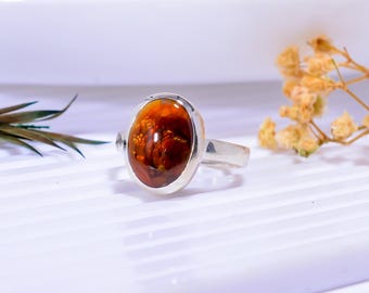 Op de afbeelding: Een zilveren ring met een ovale edelsteen in tinten oranje, bruin en rood. De edelsteen is gezet in een gepolijste zilveren band. Dit sieraad is waarschijnlijk bedoeld voor mode of versiering.