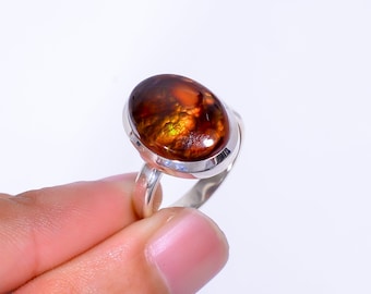 May include: A silver ring featuring an oval-shaped gemstone with a mix of brown, orange, and yellow hues. The gemstone is set in a polished silver band. The ring is held up against a white background.