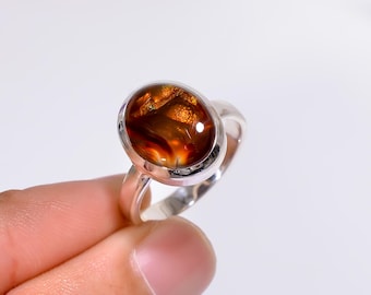 May include: A silver ring featuring an oval-shaped, cabochon-cut gemstone with a mix of brown, orange, and gold tones. The ring's band is a simple, polished silver design. The gemstone is the focal point, showcasing natural patterns.