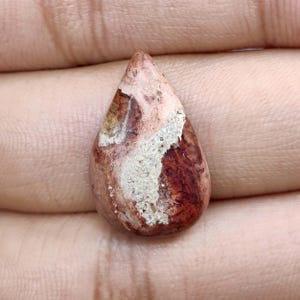 May include: A teardrop-shaped gemstone with a mix of colors. The stone features a base of deep reddish-brown, with patches of tan and white. The polished surface reflects light, highlighting the natural patterns and textures within the stone.