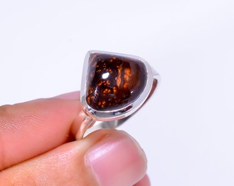 May include: A silver ring featuring a large, polished gemstone with a unique pattern of dark brown and reddish-brown hues. The stone is set in a smooth, bezel setting, creating a modern and elegant look. The ring is held by a person.