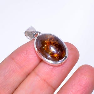 May include: Oval-shaped pendant with a silver-colored bail. The pendant features a polished, brown gemstone with golden inclusions. The gemstone is set in a silver bezel. The pendant is held by a person's fingers.