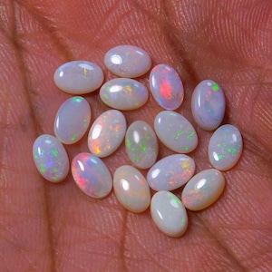 Può includere: Una collezione di opali bianchi ovali con bagliori iridescenti arcobaleno. Gli opali sono disposti su una mano.