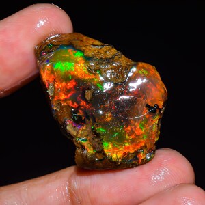 Op de afbeelding: Een ruwe, ongeslepen opaal edelsteen met levendige kleuren. De steen toont een mix van vurige roodtinten, oranje, groen en blauw, met een donkere, getextureerde matrix. De opaal wordt tussen vingers gehouden, wat de natuurlijke schoonheid benadrukt.