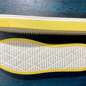 May include: Two yellow and white shoe soles are displayed against a dark blue background. The soles have a textured, diamond-patterned design on the bottom and a thick yellow border. The top sole is elevated, showing the side profile.