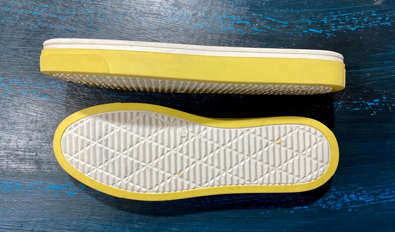 May include: Two yellow and white shoe soles are displayed against a dark blue background. The soles have a textured, diamond-patterned design on the bottom and a thick yellow border. The top sole is elevated, showing the side profile.