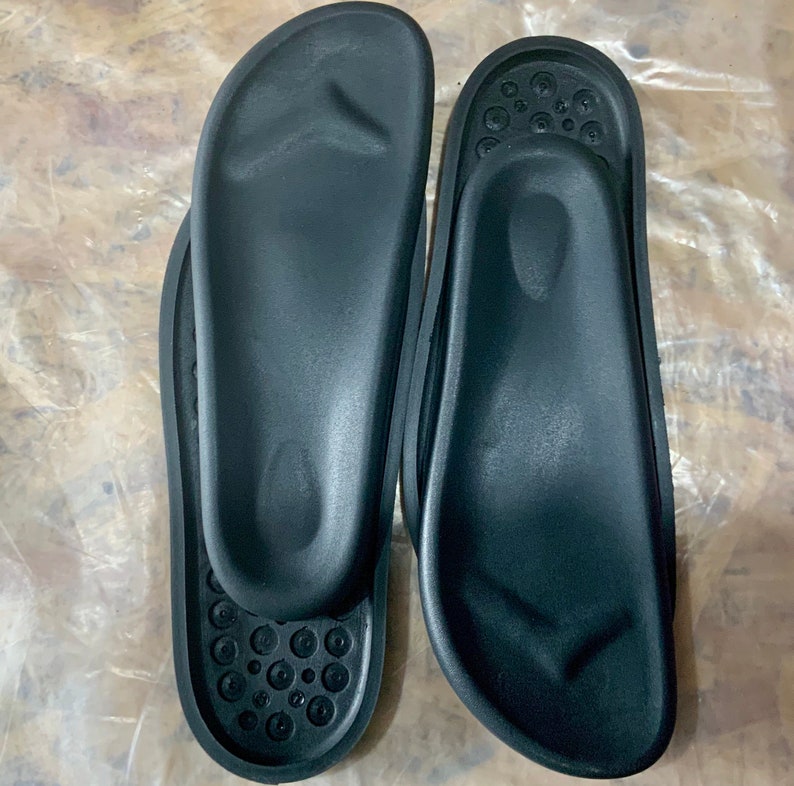 May include: Two black orthopedic shoe inserts with a textured surface. The inserts have a raised arch support and a contoured heel.