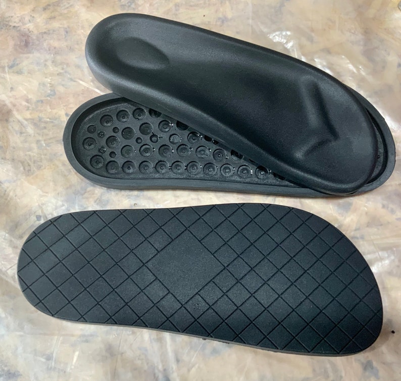 May include: Two black rubber soles for sandals. One sole has a textured surface with small circles. The other sole has a diamond pattern.