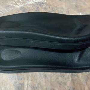 May include: Two black foam armrests for a chair or sofa. The armrests are oval shaped and have a smooth, slightly textured surface.