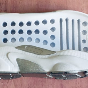 May include: A pair of white shoe soles with a silver accent. The top sole has a pattern of small holes and five horizontal lines. The bottom sole has a textured pattern.