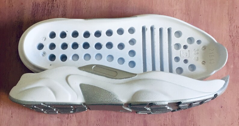 May include: A pair of white shoe soles with a silver accent. The top sole has a pattern of small holes and five horizontal lines. The bottom sole has a textured pattern.