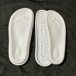 May include: A pair of white flip-flop sandals with a textured footbed. The sandals are made of a soft, flexible material and have a simple, classic design.
