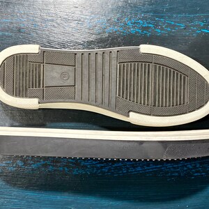May include: Two black and white sneaker soles. The bottom sole is black with a white border and a textured pattern. The top sole is also black with a white border and a textured pattern. The soles are on a blue surface.