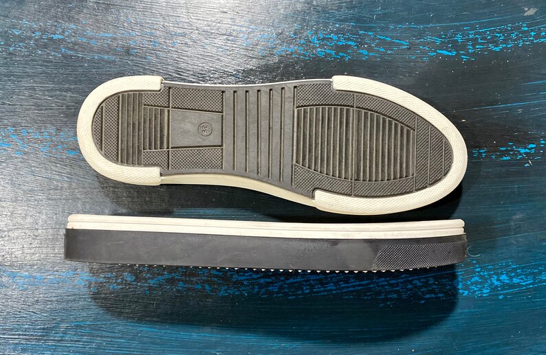 May include: Two black and white sneaker soles. The bottom sole is black with a white border and a textured pattern. The top sole is also black with a white border and a textured pattern. The soles are on a blue surface.