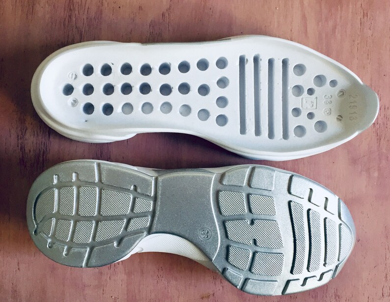 May include: A pair of white shoe soles, one with a textured rubber tread and the other with a smooth surface and a pattern of small holes. The sole with the holes has the text "38 1/3" printed on it.
