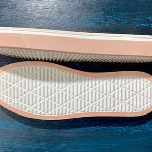 May include: Two shoe soles, one above the other, are displayed. The soles are a light pink color with a white, textured bottom. The top sole is viewed from the side, showing the pink edge and white sole. The bottom sole is viewed from the bottom.
