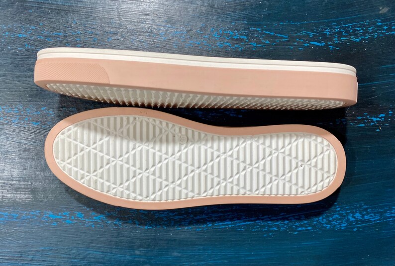 May include: Two shoe soles, one above the other, are displayed. The soles are a light pink color with a white, textured bottom. The top sole is viewed from the side, showing the pink edge and white sole. The bottom sole is viewed from the bottom.