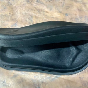 May include: Two black foam footbeds for sandals or slippers. The footbeds are curved and have a raised edge around the perimeter.