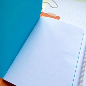 Op de afbeelding: Open notitieboek met een turquoise omslag en pagina's met stippenraster. Een lichtpaarse en wit gestreepte potlood ligt op een pagina met ruitjespapier. Een stuk bruin papier met plakband en een gouden touwtje zijn ook zichtbaar.