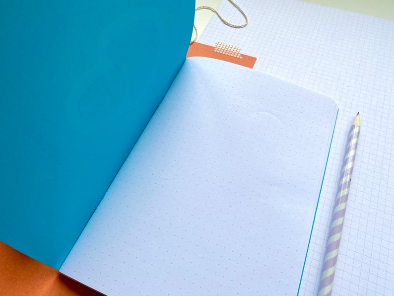 Op de afbeelding: Open notitieboek met een turquoise omslag en pagina's met stippenraster. Een lichtpaarse en wit gestreepte potlood ligt op een pagina met ruitjespapier. Een stuk bruin papier met plakband en een gouden touwtje zijn ook zichtbaar.