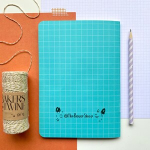 Op de afbeelding: Een turquoise notitieboek met een rasterpatroon, een potlood met paarse en witte strepen en een rol bakkersgaren met gouden accenten. Het notitieboek heeft een paddenstoelafbeelding en de tekst "@TheRoverShop".