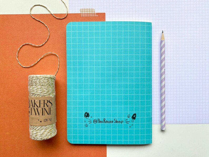 Op de afbeelding: Een turquoise notitieboek met een rasterpatroon, een potlood met paarse en witte strepen en een rol bakkersgaren met gouden accenten. Het notitieboek heeft een paddenstoelafbeelding en de tekst "@TheRoverShop".