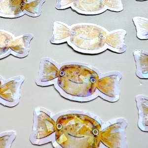 May include: A set of ten colorful, iridescent stickers featuring a cartoon fish shaped like a candy. The fish have yellow bodies with brown spots and black eyes. The stickers are perfect for adding a whimsical touch to laptops, water bottles, or journals.