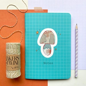 Op de afbeelding: Een turquoise notitieboek met een rasterpatroon, een paddenstoelsticker en het woord 'Notes'. Een rol goud-wit bakkersgaren en een gestreepte potlood zijn ook aanwezig. De achtergrond is oranje en wit.
