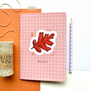 Op de afbeelding: Een roze notitieboek met een rasterpatroon en het woord "Notes", met een rode bladsticker. Een gestreepte potlood en een rol bakkersgaren zijn ook aanwezig. Het garen is gelabeld "Baker's Twine 120 YD".