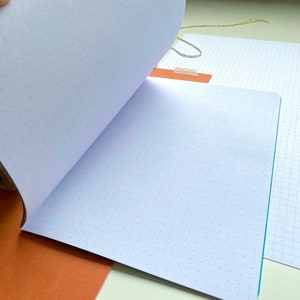 Op de afbeelding: Open notitieboek met stippenrasterpagina's, ideaal voor journaling of schetsen. Een potlood met paarse en witte strepen en een geruit papier zijn ook zichtbaar. Het notitieboek is open en onthult de lege pagina's.