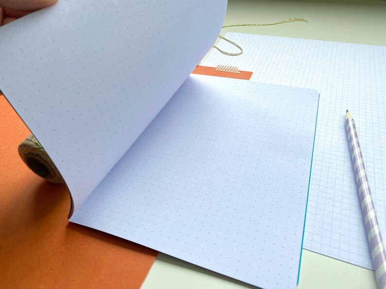Op de afbeelding: Open notitieboek met stippenrasterpagina's, ideaal voor journaling of schetsen. Een potlood met paarse en witte strepen en een geruit papier zijn ook zichtbaar. Het notitieboek is open en onthult de lege pagina's.