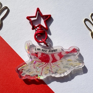 May include: A red star-shaped keychain with a clear charm featuring a red and white shrimp with the text "I AM JUST A BABY..."