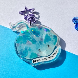 Può includere: Un portachiavi in resina blu e bianca a forma di razza con glitter incorporati nella resina. Il portachiavi ha un ciondolo a forma di stella viola e un anello diviso argentato. La razza ha un viso con due occhi e una bocca. Il testo "Brain not Braining..." è scritto sul fondo del portachiavi.
