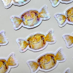 May include: A set of nine iridescent stickers featuring cartoon pufferfish with candy-like tails. The fish are yellow, orange, and white with black eyes and a smiling mouth.