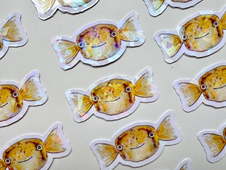 May include: A set of nine iridescent stickers featuring cartoon pufferfish with candy-like tails. The fish are yellow, orange, and white with black eyes and a smiling mouth.
