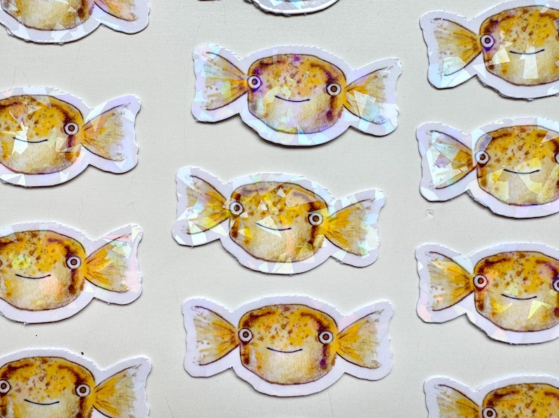 May include: A set of 10 iridescent stickers featuring a cartoon pufferfish with a candy-like design. The stickers are white with a holographic finish and feature a yellow and brown pufferfish with black eyes and a smile.