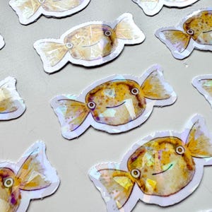 May include: A set of ten iridescent stickers featuring a cartoon fish shaped like a candy. The fish are yellow and brown with black eyes and a white outline.