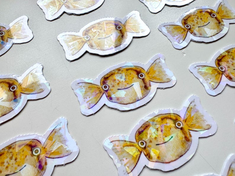 May include: A set of ten iridescent stickers featuring a cartoon fish shaped like a candy. The fish are yellow and brown with black eyes and a white outline.